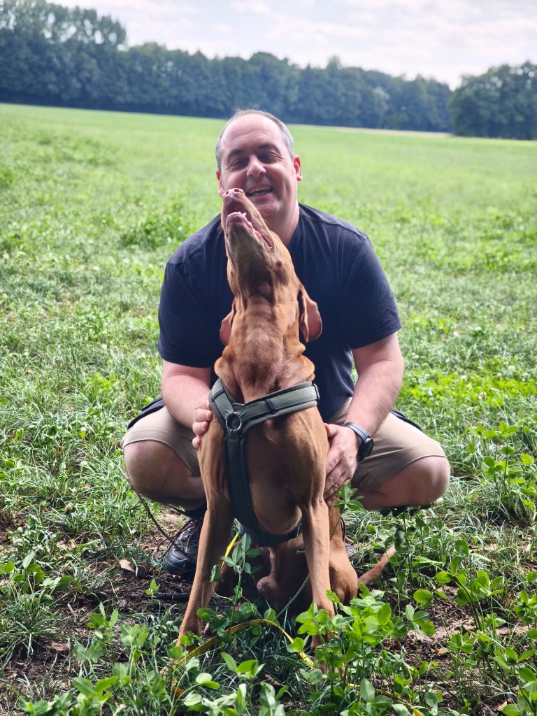 Mann kniet in einer Wiese und interagiert fröhlich mit einem Hund.