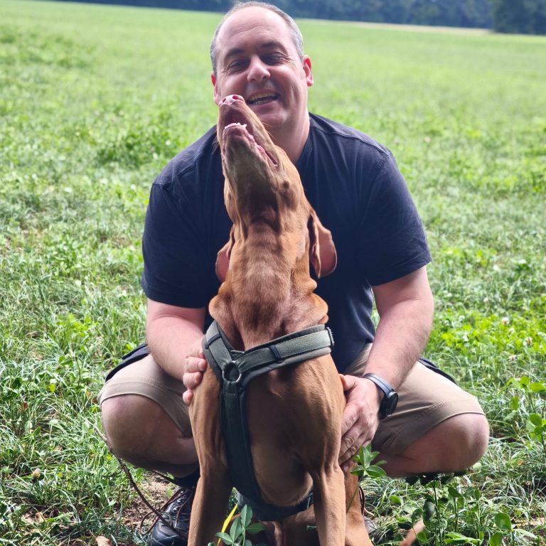 Mann kniet in einer Wiese und interagiert fröhlich mit einem Hund.